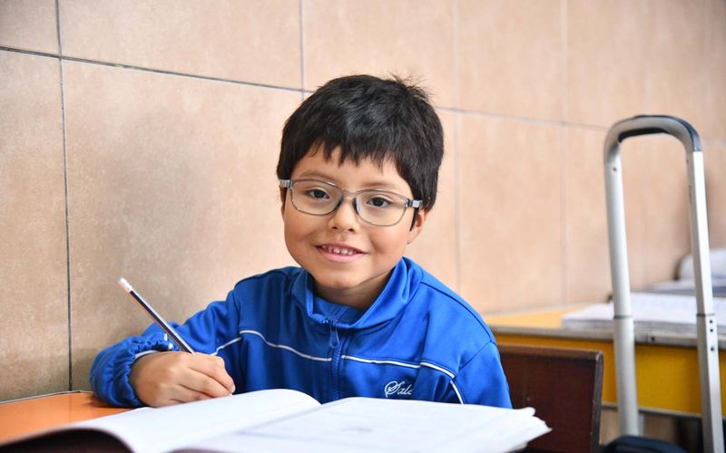 Niño de primaria con lentes escribiendo en su cuaderno y sonriendo en el salón