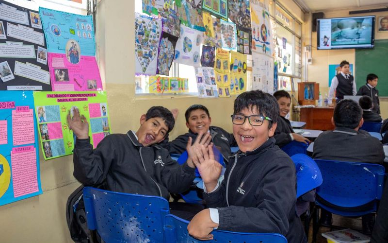 Alumnos saludando y sonriendo en el aula, rodeados de trabajos pegados en las paredes