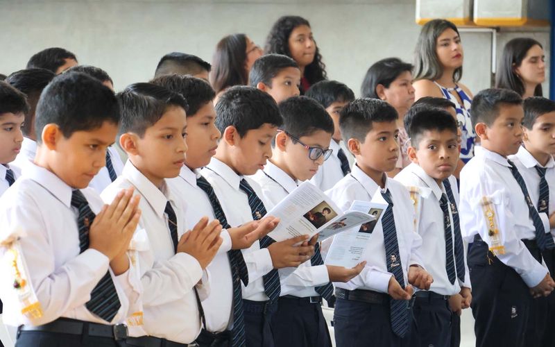 Alumnos de primaria con camisa blanca y corbata rezando con las manos juntas y el devocionario