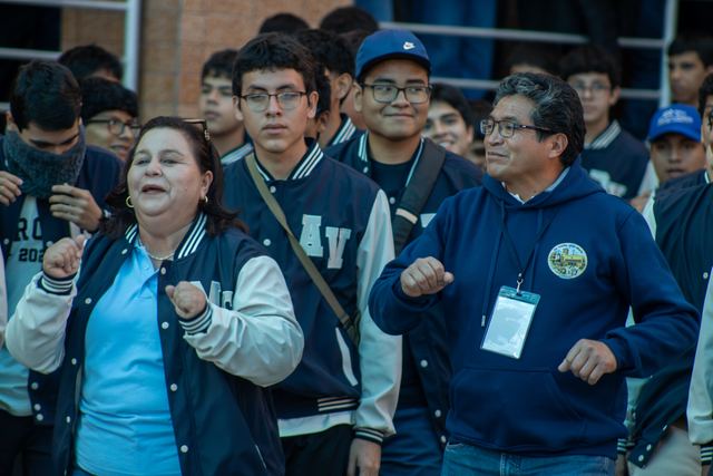 Una educadora y un docente bailan junto a estudiantes con chaquetas de promoción durante la recepción