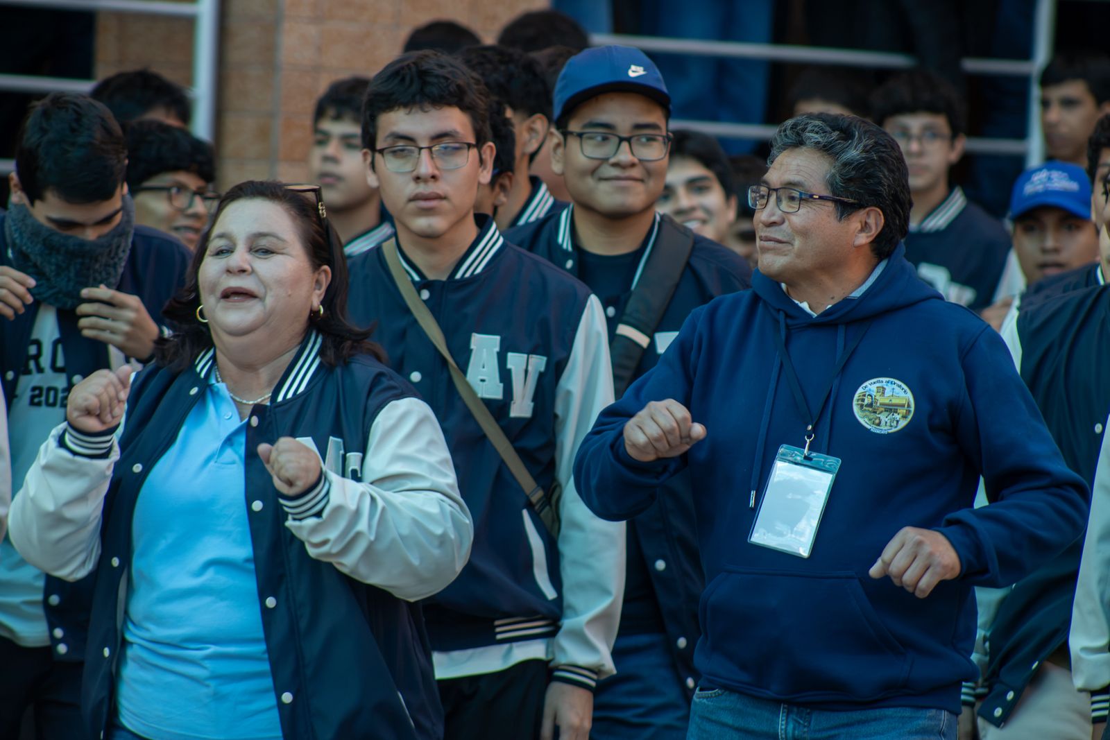 Una educadora y un docente bailan junto a estudiantes con chaquetas de promoción durante la recepción